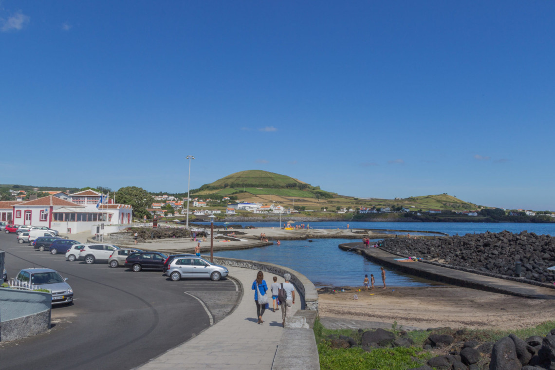 Porto Martins Natural Pools on the eastern coast of Terceira Island, with sheltered seawater swimming areas, a small beach, waterfront promenade and green volcanic hills behind the village.