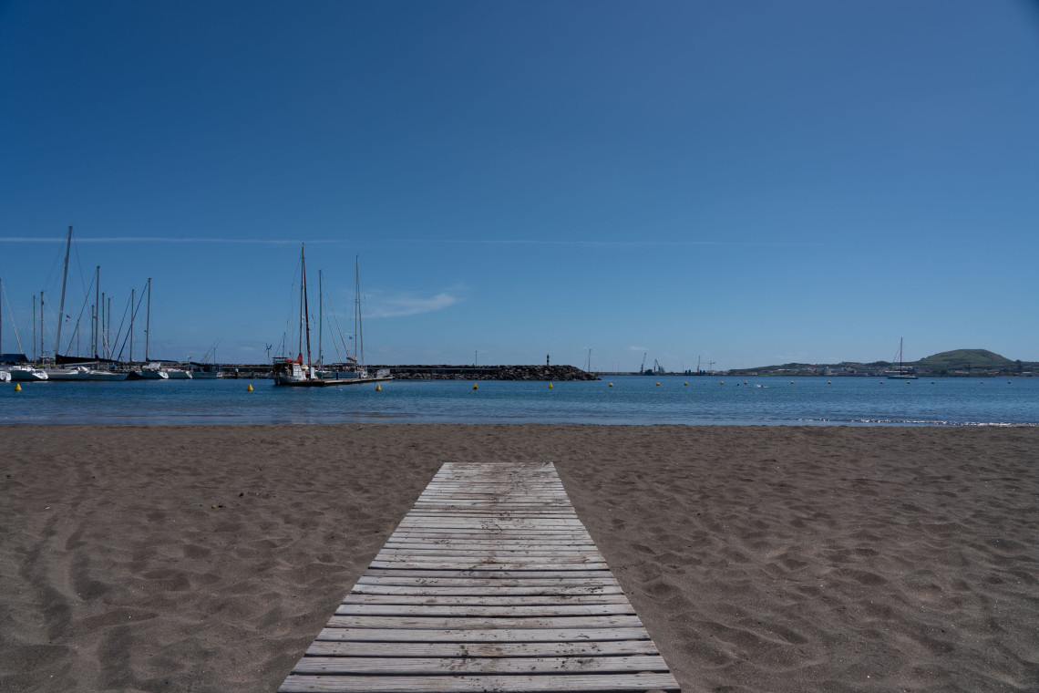 View from Praia Grande in Praia da Vit&oacute;ria on Terceira Island, with a wooden walkway crossing the dark sandy beach toward the calm Atlantic bay. Sailboats and the Praia da Vit&oacute;ria Marina are visible along the waterfront.