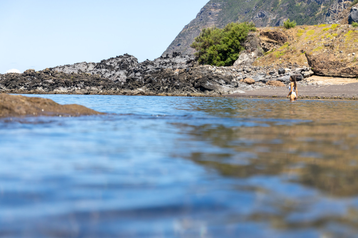 A visitor walks through the shallow water at Praia da Areia in Vila do Corvo, Corvo Island, Azores, Portugal. Dark volcanic sand, basalt rocks and steep coastal slopes frame the sheltered Atlantic beach.