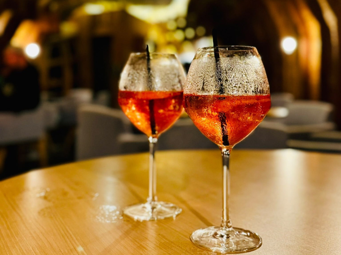 Two chilled orange-colored drinks served in wine glasses at Cella Bar in Madalena, with the restaurant&rsquo;s warm wooden interior softly visible in the background.
