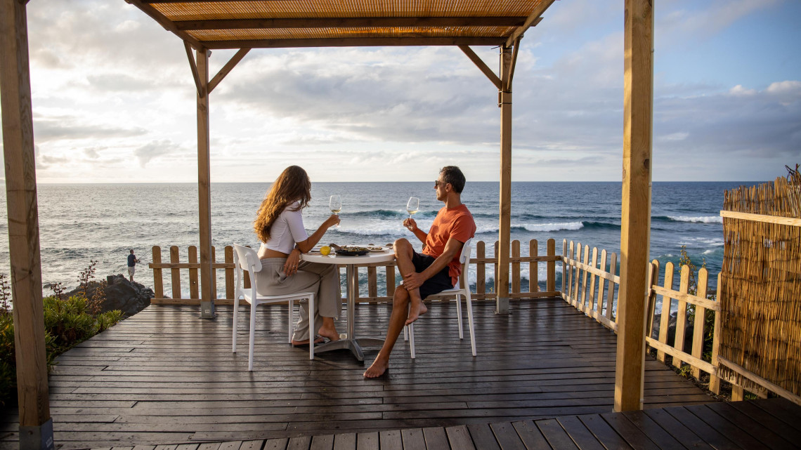 Couple enjoying an oceanfront meal and wine near Areal de Santa B&aacute;rbara in Ribeira Grande, with the Atlantic Ocean stretching behind their terrace.