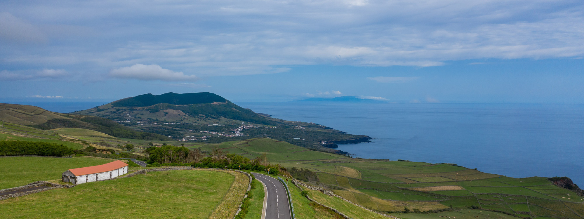 Panoramic view from Serra Branca on Graciosa Island, showing a winding road through green agricultural fields toward the Atlantic coastline. Rolling volcanic terrain and a distant Azorean island are visible beneath a cloud-covered sky.