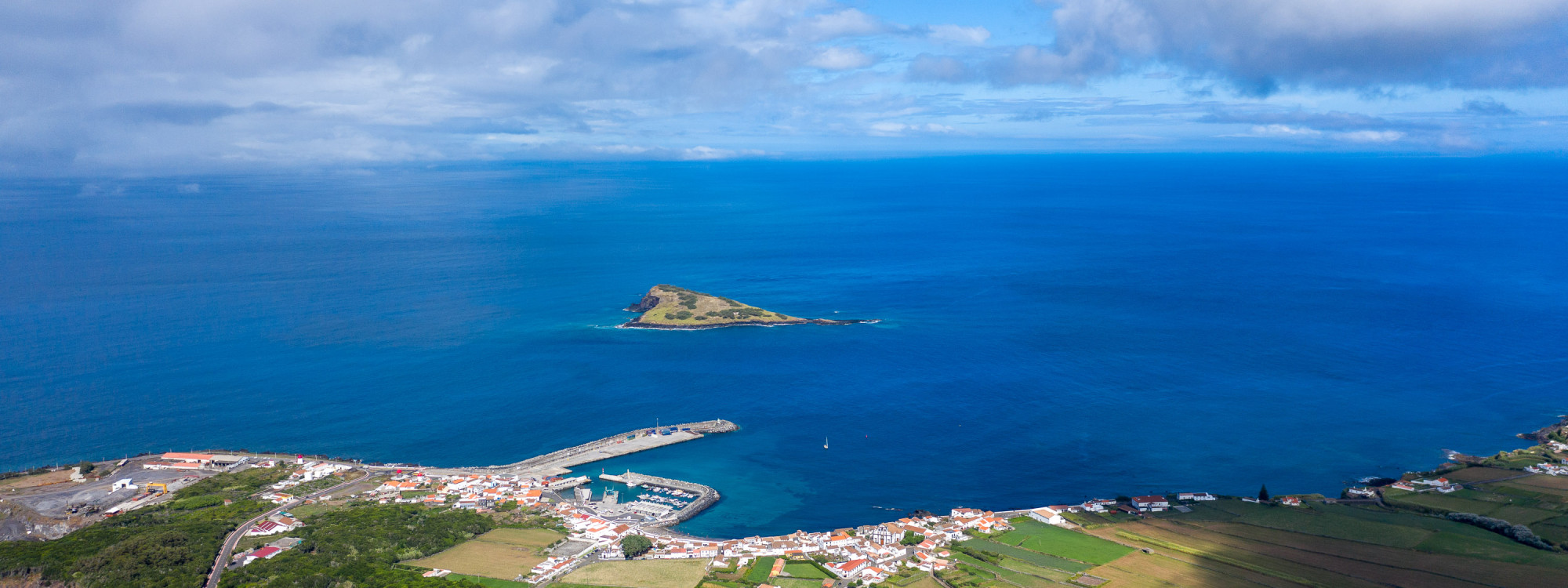Panoramic view over Vila da Praia on Graciosa Island, showing the harbor and red-roofed settlement beside the Atlantic Ocean with Ilhéu da Praia offshore.