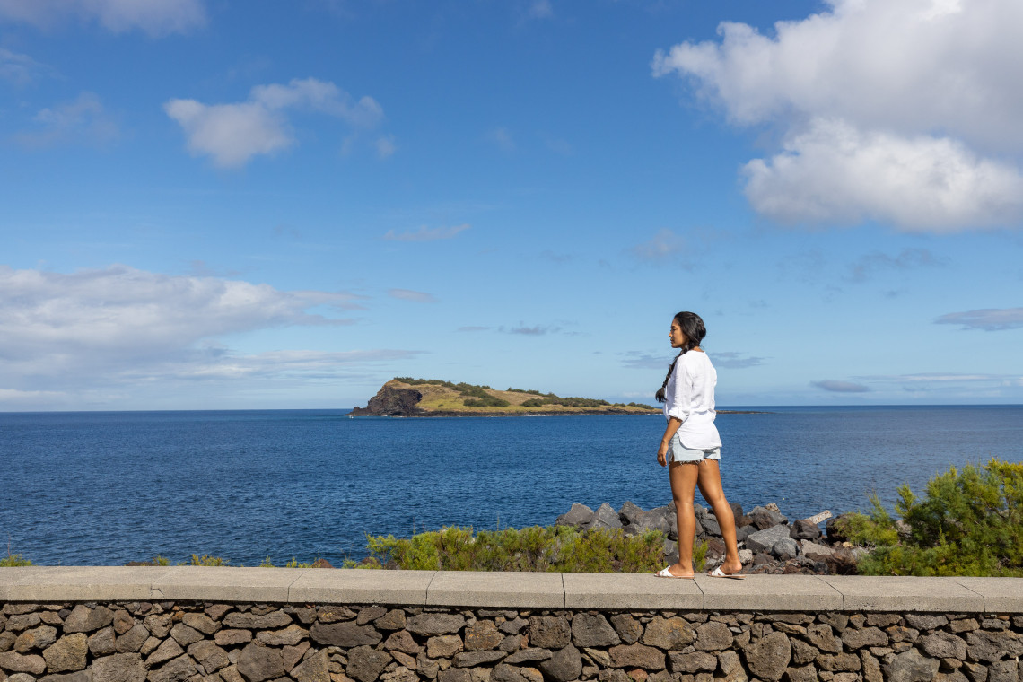 Traveler walking along the coast of S&atilde;o Mateus on Graciosa Island with Ilh&eacute;u da Praia rising from the Atlantic Ocean in the background.