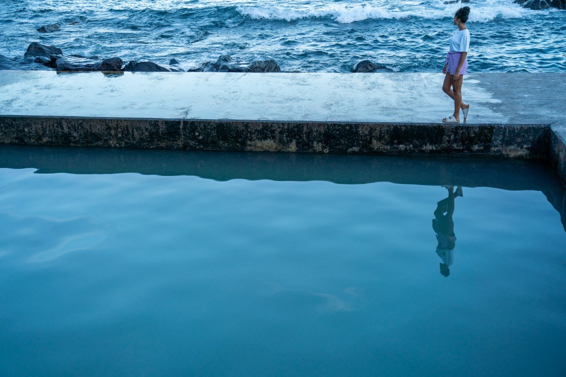Traveler walking beside a calm seawater swimming pool on the coast of Santa Cruz da Graciosa, with Atlantic waves breaking immediately beyond the concrete sea wall.