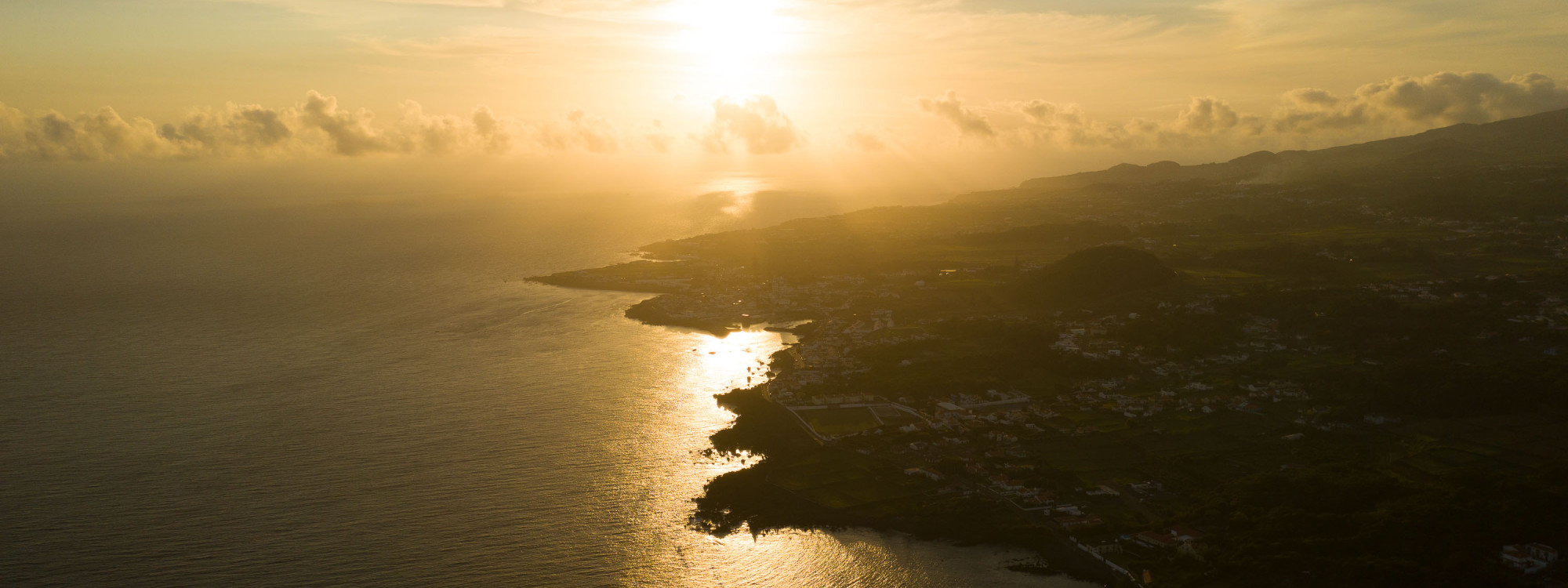 Aerial view of the Atlantic coastline near Angra do Heroísmo on Terceira Island at sunset. Golden light reflects across the ocean beside the island’s volcanic shoreline and coastal settlements.