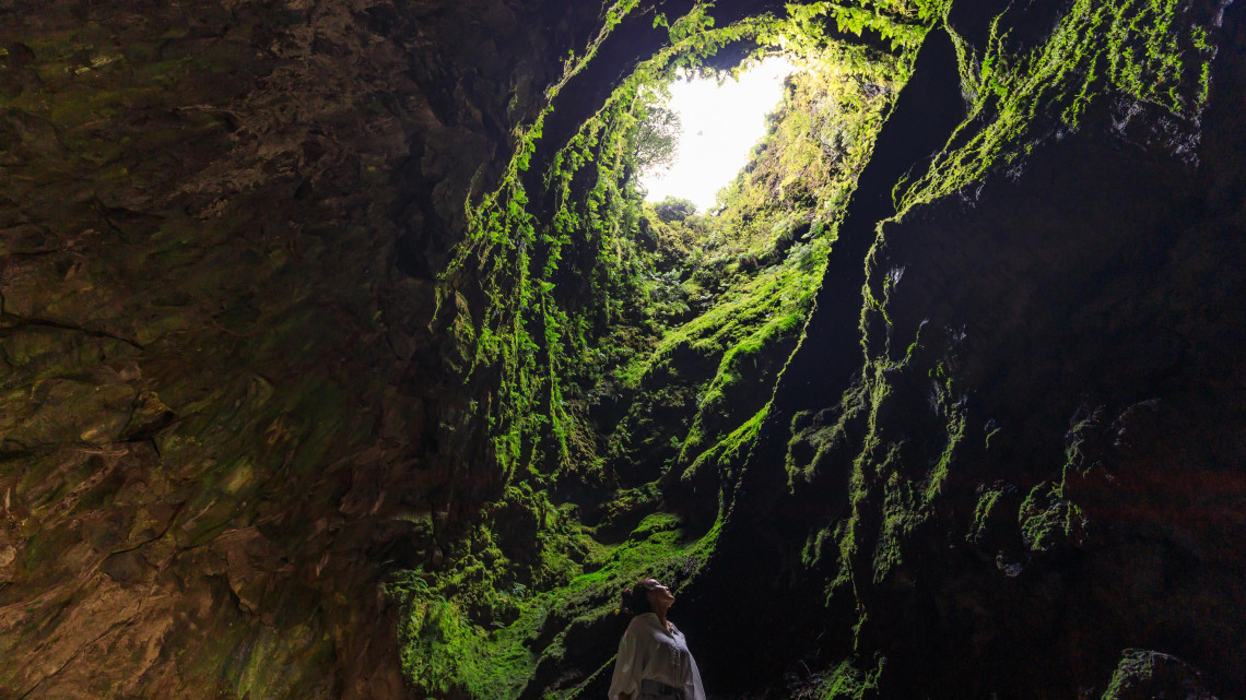 Interior of Algar do Carv&atilde;o on Terceira Island, with a visitor beneath the open volcanic chimney surrounded by moss-covered rock walls and dense vegetation.