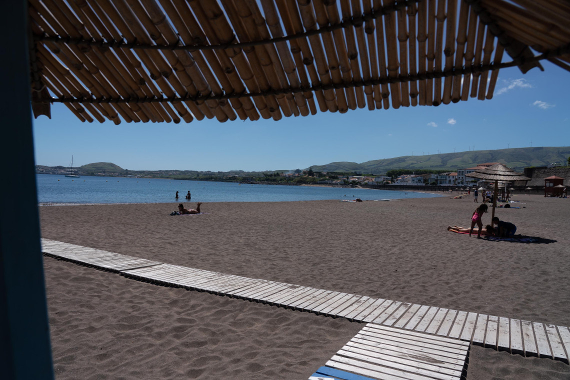 Praia Grande in Praia da Vit&oacute;ria on Terceira Island, viewed from beneath a shaded beach structure. A wooden walkway crosses the broad volcanic-sand beach beside the calm waters of the bay.