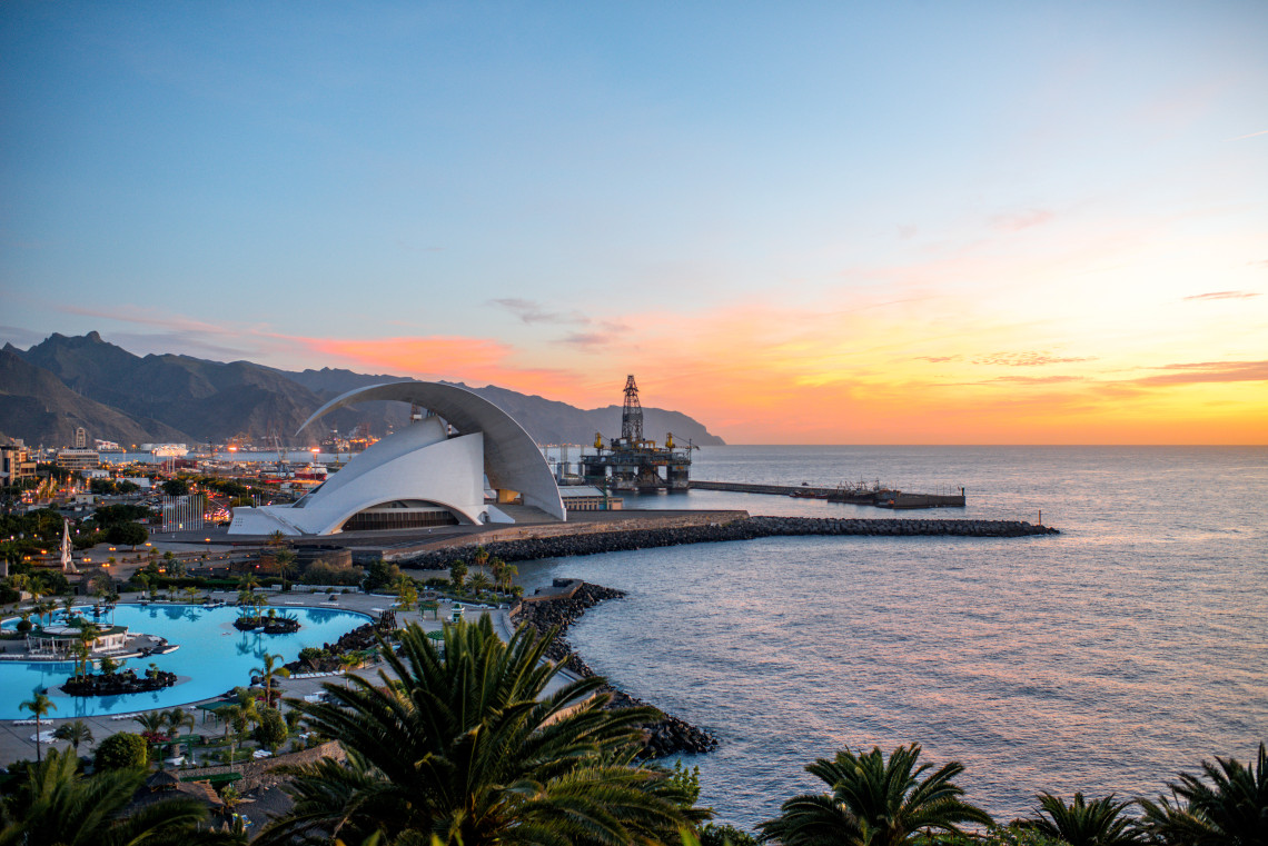 View of the Auditorio de Tenerife Ad&aacute;n Mart&iacute;n on the waterfront of Santa Cruz de Tenerife, with palm trees, the Atlantic Ocean, and warm sunset light over the coast.