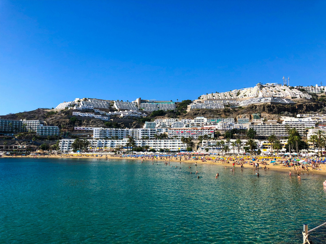 Playa de Puerto Rico on the southwest coast of Gran Canaria, with clear Atlantic waters, a sandy beach, and white resort buildings rising across the surrounding hillsides.