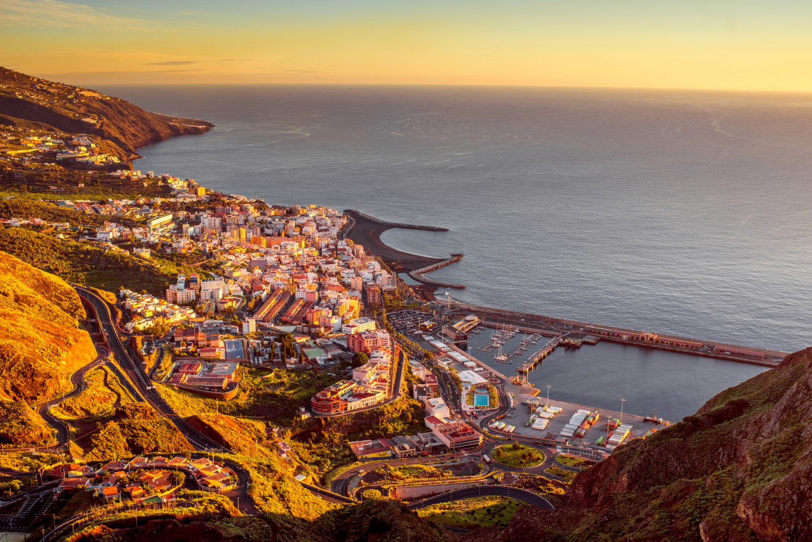 Elevated view of Santa Cruz de La Palma at sunset, showing the compact coastal city, harbour, winding roads, and steep volcanic slopes beside the Atlantic Ocean.