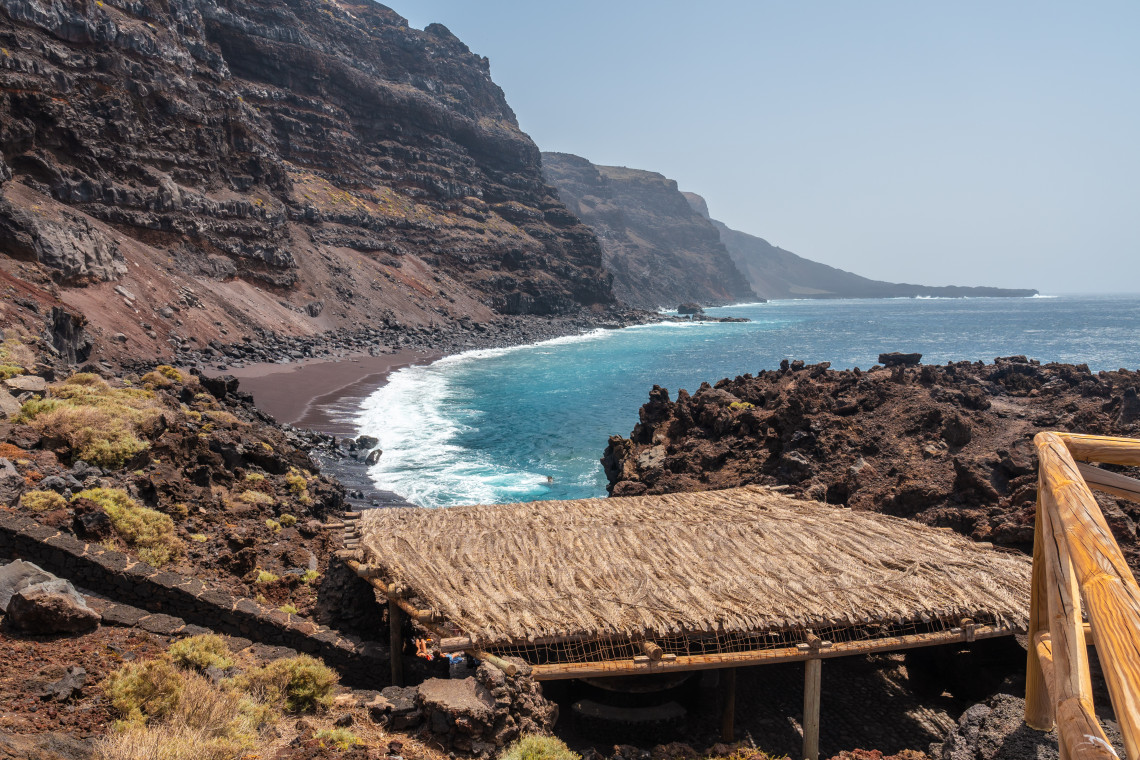 Playa del Verodal on El Hierro, with dark volcanic sand, turquoise Atlantic water, rugged coastal cliffs, and a rustic shelter overlooking the beach.