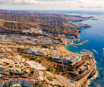 Aerial view of Puerto Rico on the southwest coast of Gran Canaria, showing its marina, sheltered bay, resort buildings, and rugged volcanic hills beside the Atlantic Ocean.