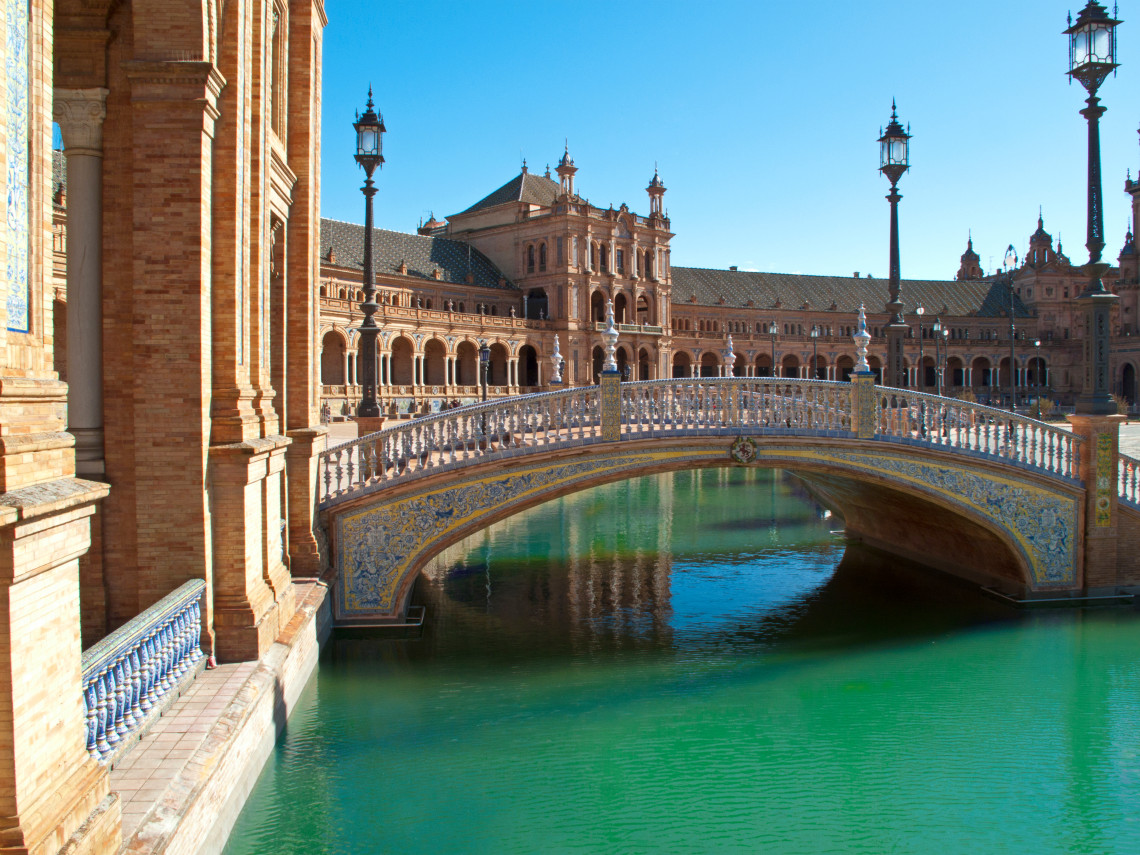 An arched bridge decorated with blue and yellow ceramic tiles crosses the canal at Plaza de Espa&ntilde;a in Seville, Spain. The brick galleries and towers of the landmark rise behind the turquoise water.