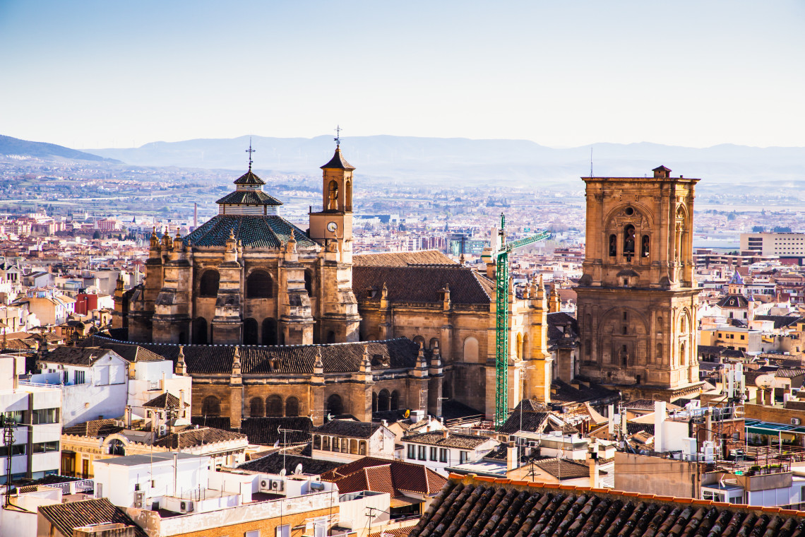 Granada Cathedral rises above the tiled rooftops of central Granada, Spain, with its dome, bell tower and stone fa&ccedil;ades visible against the surrounding city and distant mountains.