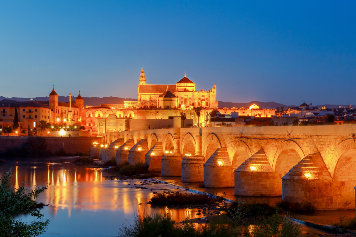 C&oacute;rdoba&rsquo;s Roman Bridge crosses the Guadalquivir River toward the illuminated Mosque-Cathedral and historic centre of C&oacute;rdoba, Spain. Lights beneath the stone arches reflect across the river at dusk.