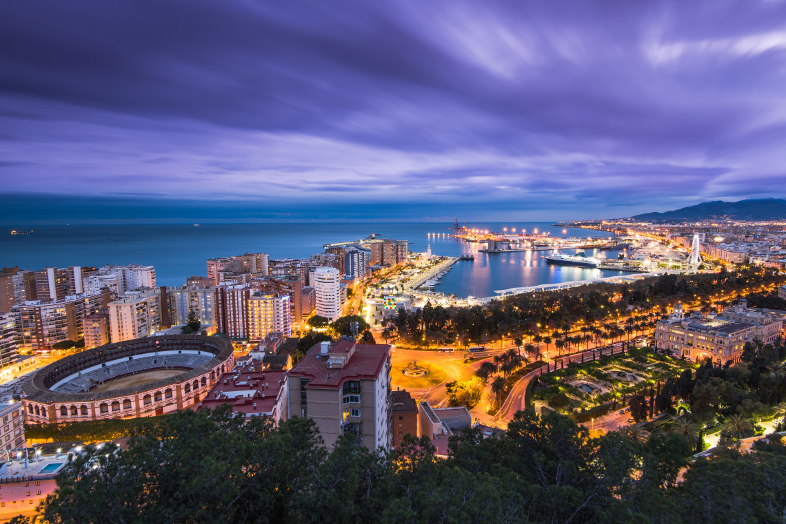 An elevated evening view from Mirador de Gibralfaro shows M&aacute;laga, Spain, including La Malagueta bullring, the waterfront, harbour and illuminated city streets beside the Mediterranean Sea.