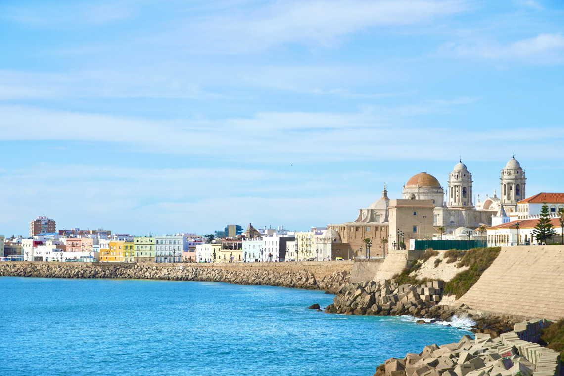 C&aacute;diz Cathedral stands above the Campo del Sur waterfront in C&aacute;diz, Spain, with its golden dome and twin towers overlooking the Atlantic Ocean. Colourful buildings extend along the fortified shoreline.