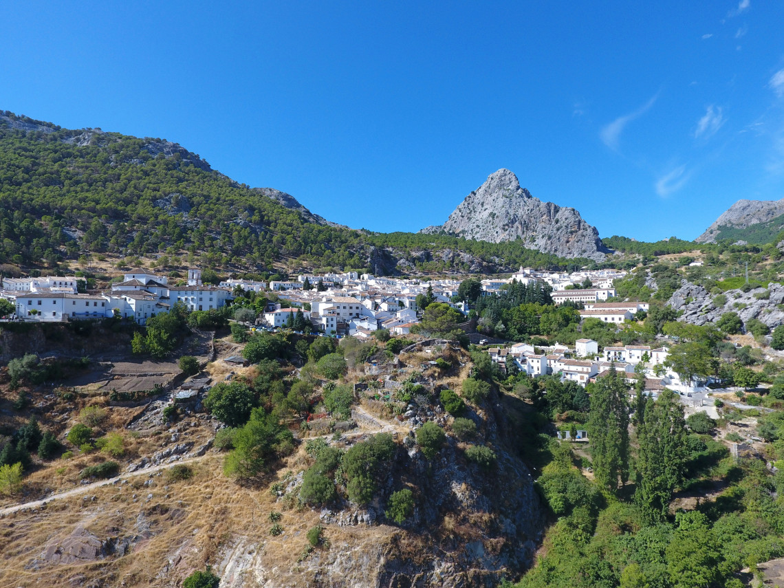 Whitewashed houses spread across the hills beneath a prominent limestone peak in Grazalema, Spain. Woodland and rocky slopes surround the village in the Sierra de Grazalema landscape.
