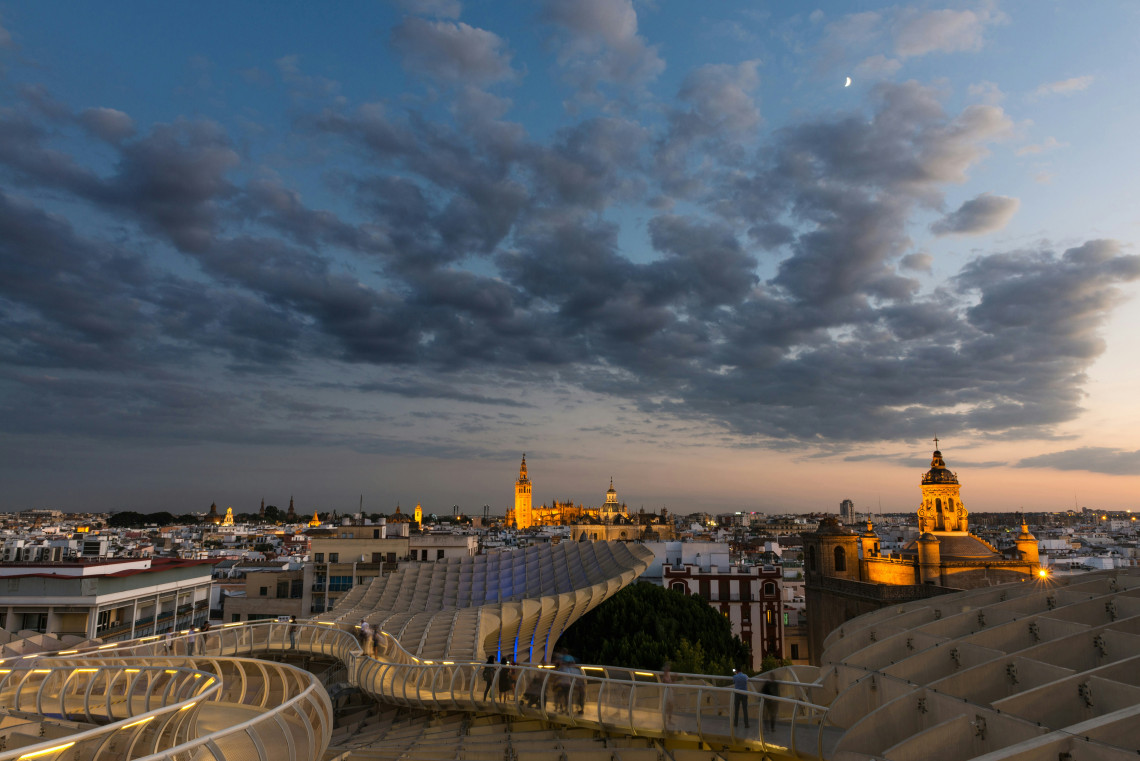 An elevated twilight view from Setas de Sevilla looks across the curved wooden walkways of Metropol Parasol toward the historic skyline of Seville, Spain. Illuminated church towers rise beneath dramatic evening clouds.