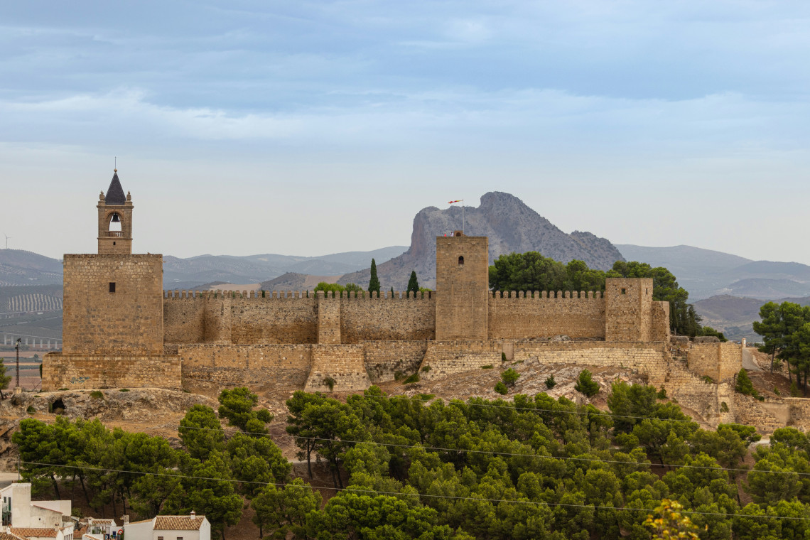 The stone walls and defensive towers of the Alcazaba of Antequera stand above Antequera, Spain. Pe&ntilde;a de los Enamorados rises behind the fortress across the Andalusian landscape.