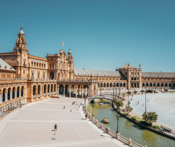 Panoramic view of Plaza de España in Seville, Spain, showing its curved brick pavilion, central canal, bridges and open pedestrian square beneath a clear blue sky.