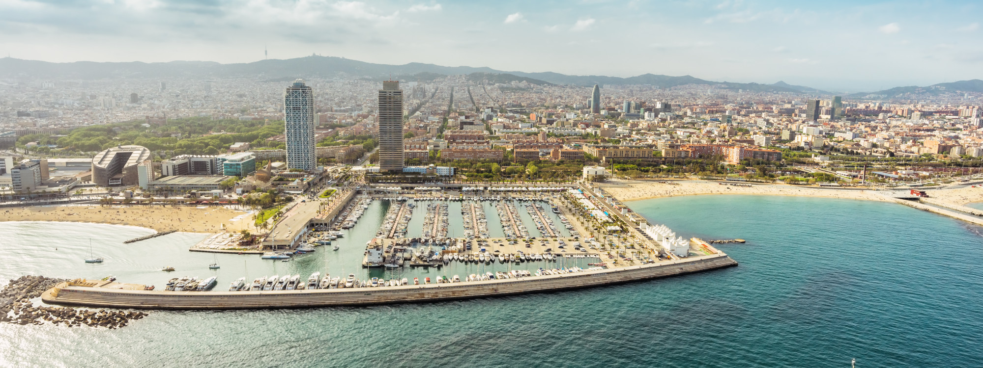 Aerial view of Port Olímpic in Barcelona, Spain, showing the marina, Mediterranean beaches and the two waterfront towers. The city extends inland towards the surrounding hills.