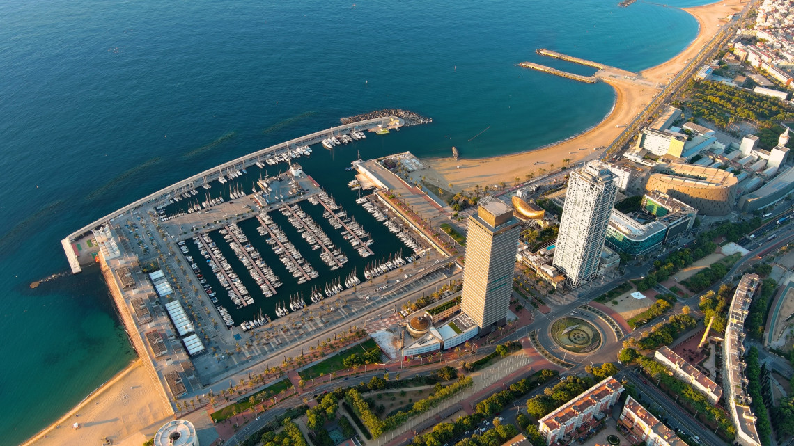 Aerial view of Port Ol&iacute;mpic in Barcelona, Spain, with rows of boats inside the marina and sandy beaches extending along the Mediterranean coast. The waterfront towers and surrounding avenues frame the harbour.
