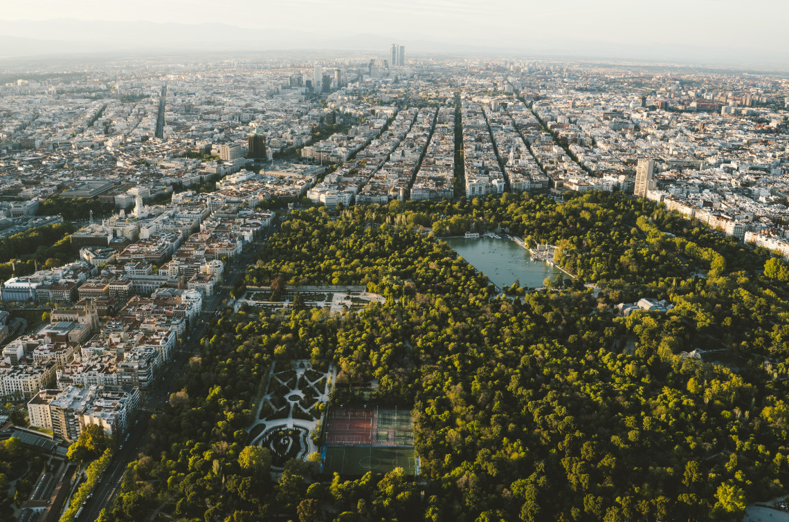 Aerial view of El Retiro Park in Madrid, Spain, showing its wooded grounds, formal gardens and central boating lake. Dense city neighbourhoods and long avenues extend around the park.