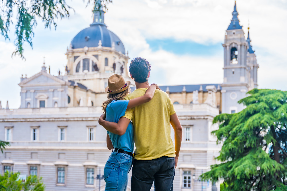 A couple embraces while looking towards Almudena Cathedral in Madrid, Spain. The cathedral&rsquo;s domed roof, pale stone fa&ccedil;ade and bell tower rise behind them.