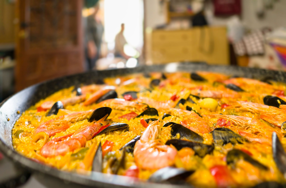 A large pan of seafood paella in Valencia, Spain, is prepared with saffron rice, prawns, mussels and vegetables. The traditional Spanish dish fills the foreground of the restaurant setting.