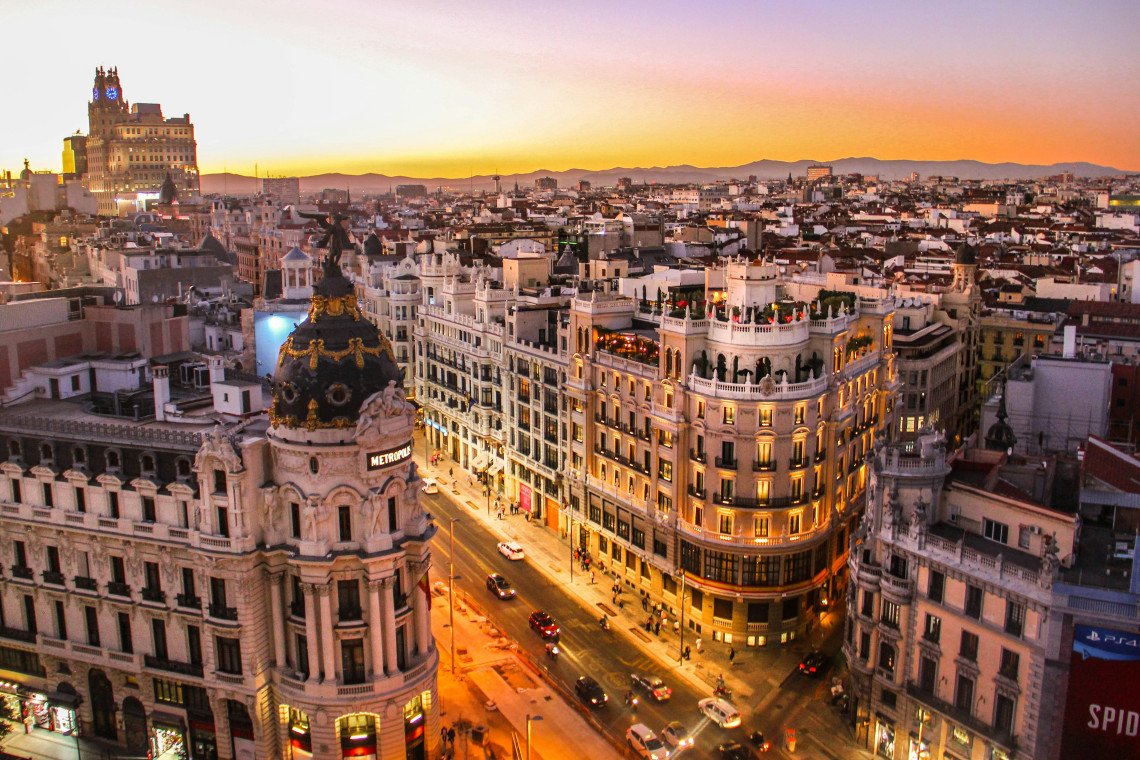 Elevated sunset view of Gran V&iacute;a in Madrid, Spain, with the domed Metr&oacute;polis Building overlooking the illuminated avenue. Historic fa&ccedil;ades, traffic and rooftops extend towards the surrounding hills.