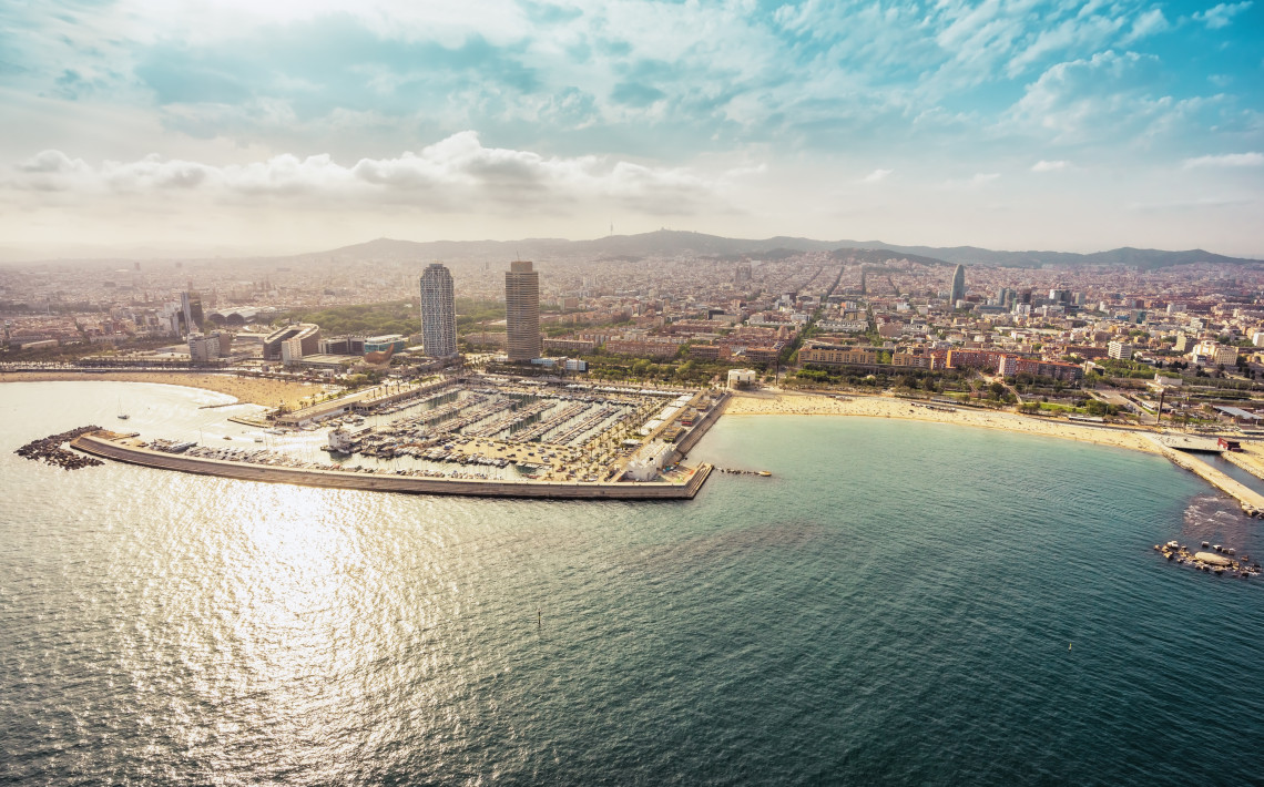 Panoramic aerial view of Port Ol&iacute;mpic in Barcelona, Spain, showing the marina, waterfront towers and beaches beside the Mediterranean Sea. Barcelona&rsquo;s urban districts extend inland towards the hills.