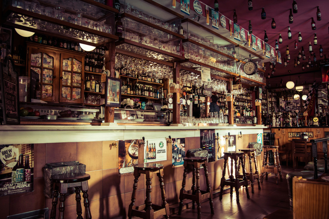 A traditional pub interior in Madrid, Spain, features a long wooden counter, bar stools and shelves filled with bottles and glassware. Beer taps, posters and hanging decorations cover the warmly lit space.