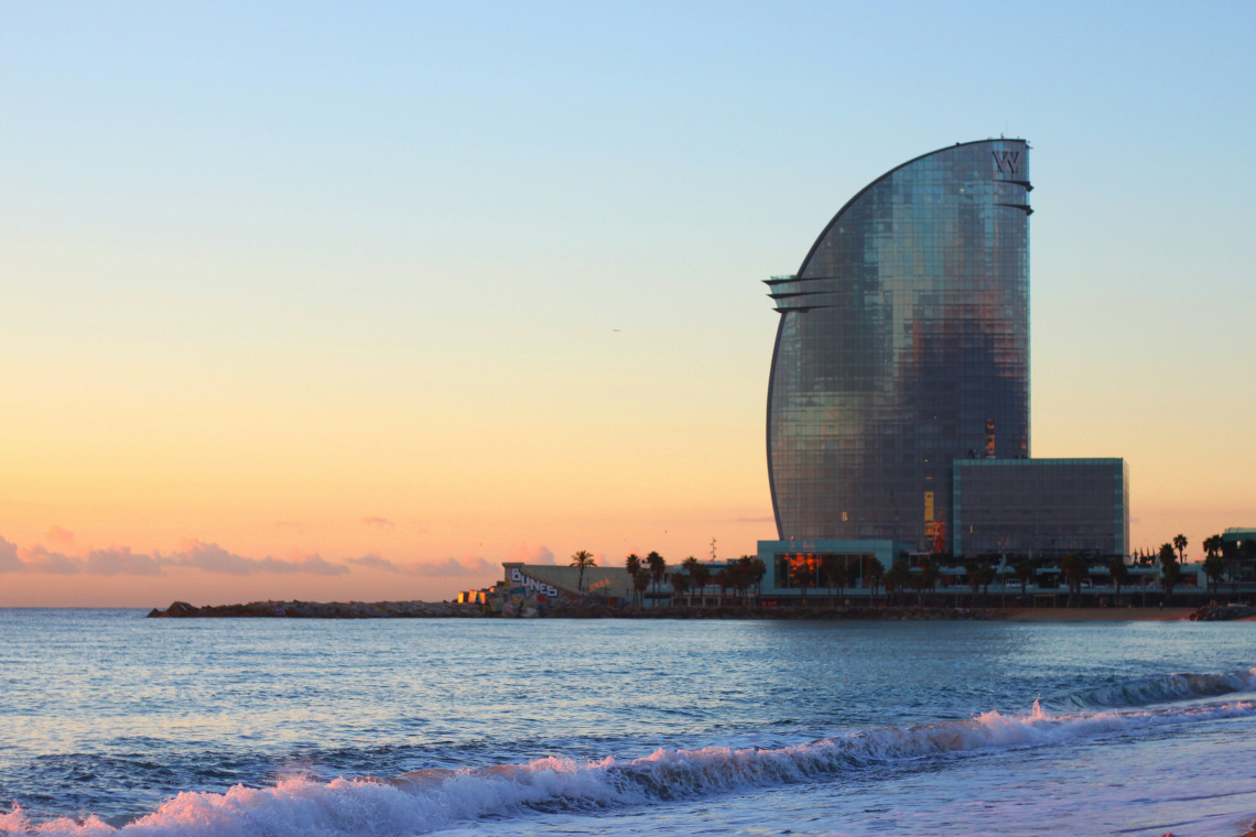The sail-shaped W Barcelona hotel rises beside Barceloneta Beach in Barcelona, Spain, during sunset. Waves reach the sandy Mediterranean shoreline beneath a pale orange sky.