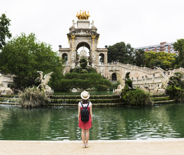 A traveller stands beside the lake facing the Cascada Monumental in Parc de la Ciutadella, Barcelona, Spain. The ornamental fountain, stone sculptures and landscaped greenery rise behind the water.