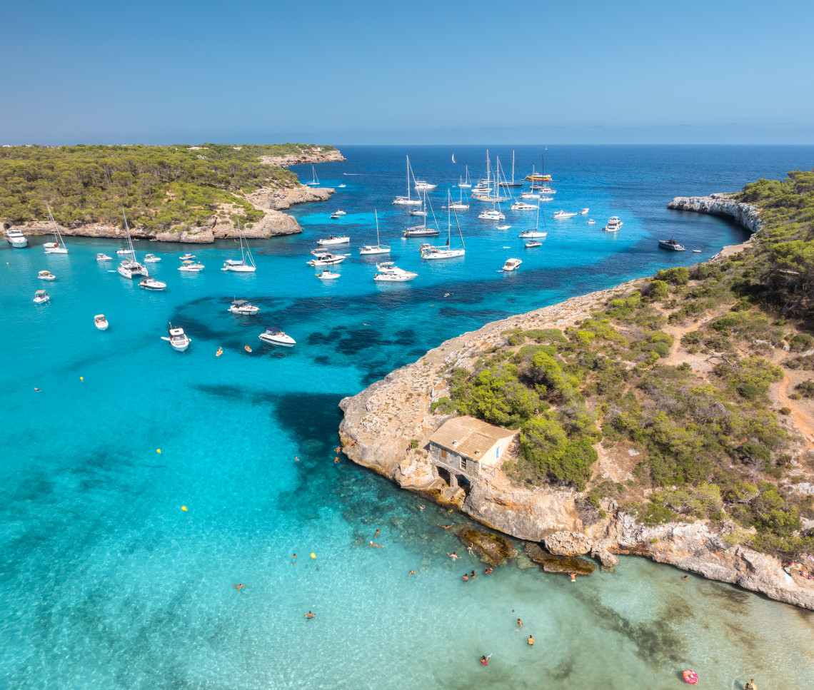 Aerial view of a turquoise cove in Mallorca with anchored sailboats and swimmers near the shore