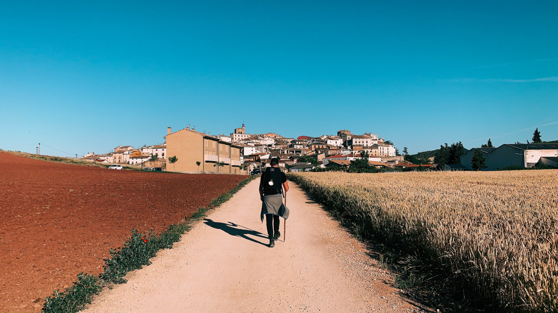 A solo pilgrim walks toward a hilltop village along a broad Camino Franc&eacute;s trail in Navarre, Spain. Red farmland and a mature wheat field border the path beneath a clear blue sky.