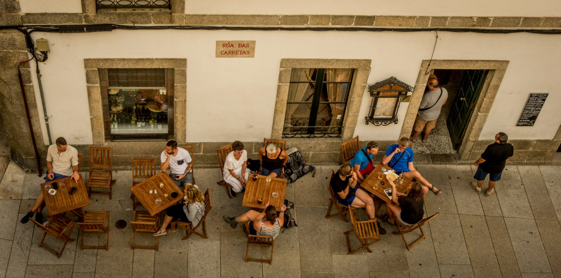 Visitors sit at wooden caf&eacute; tables along R&uacute;a das Carretas in the historic centre of Santiago de Compostela, Spain. The overhead view shows the stone-paved street and traditional building fa&ccedil;ades near the cathedral.