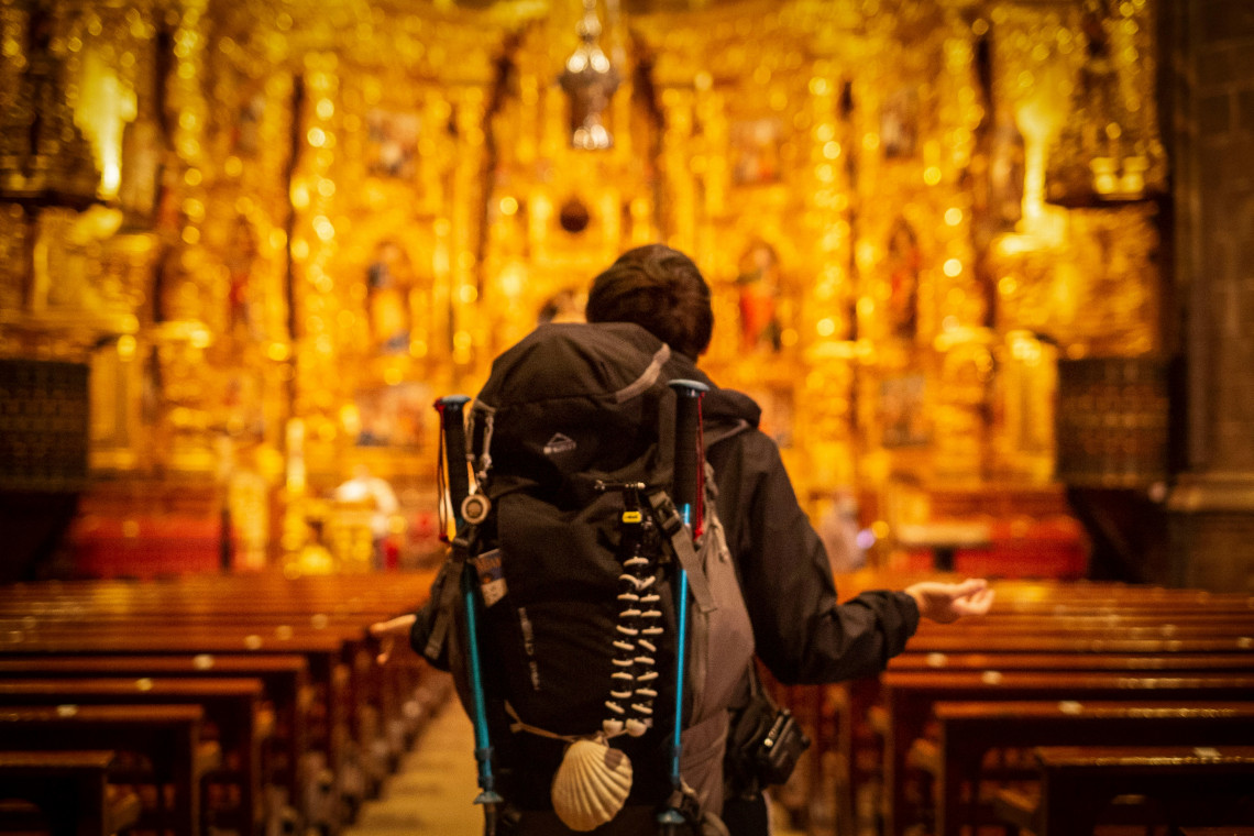 A Camino de Santiago pilgrim carrying trekking poles and a backpack decorated with a scallop shell stands before an elaborate gilded church altar in Spain. Wooden pews lead toward the brightly illuminated Baroque interior.