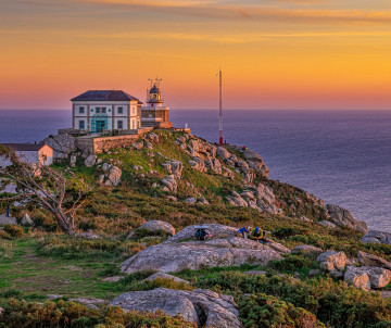 Cape Fisterra Lighthouse stands on a rocky Atlantic headland in Fisterra, Galicia, as the sky turns orange at sunset. Pilgrims gather among the granite rocks near the traditional final destination of the Camino de Santiago.