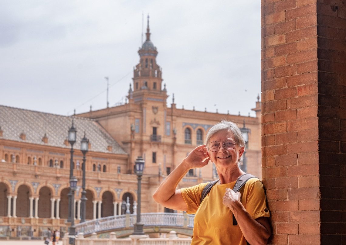 A smiling senior traveller stands beneath a brick archway at Plaza de Espa&ntilde;a in Seville, Spain. The landmark&rsquo;s tiled bridges, columns and central tower are visible behind her.