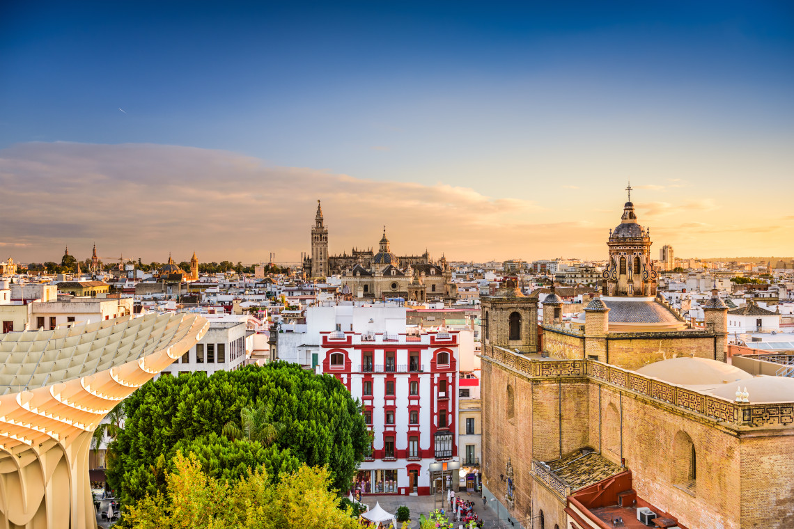 Elevated view across the historic centre of Seville, Spain, with La Giralda and Seville Cathedral rising above white buildings. The Metropol Parasol and surrounding church towers frame the cityscape at sunset.