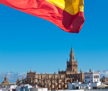 The Spanish flag waves above Seville Cathedral and the La Giralda bell tower in Seville, Spain. The historic landmark rises against a clear blue sky beyond the city’s whitewashed buildings.