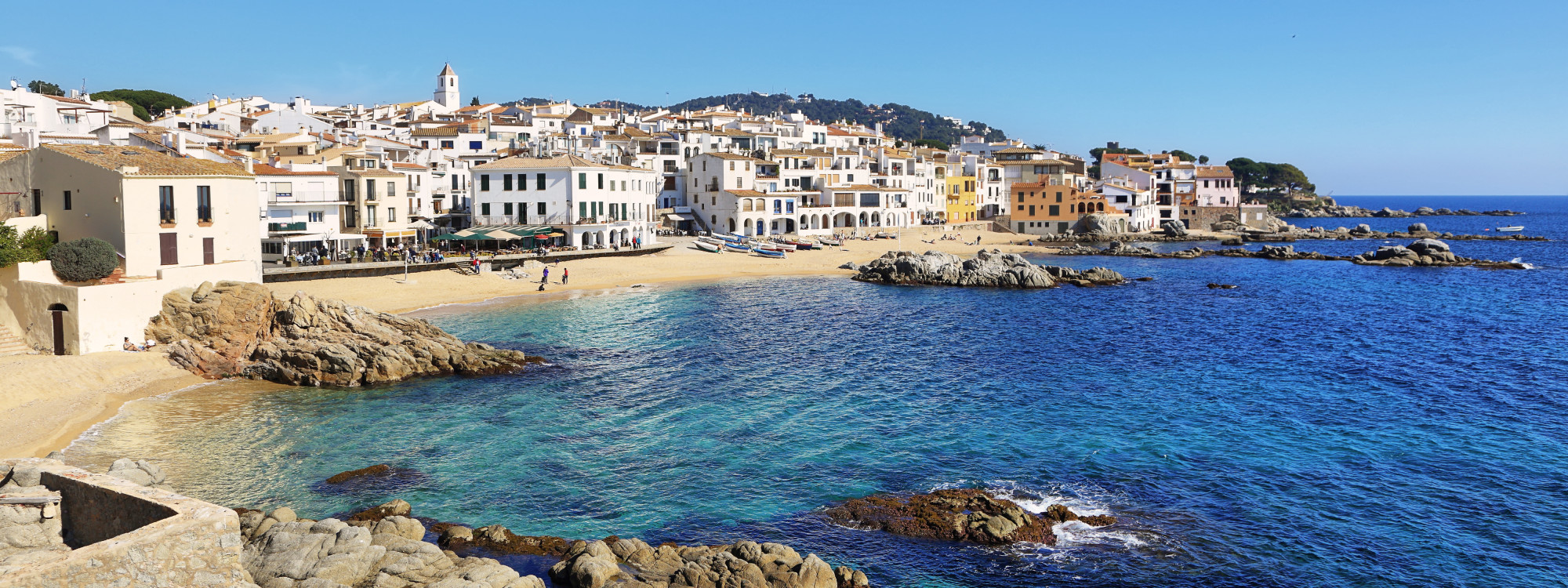 White buildings line the rocky Mediterranean waterfront of Calella de Palafrugell in Palafrugell, Spain. Clear blue water surrounds the small beaches and coves of this Costa Brava fishing village.