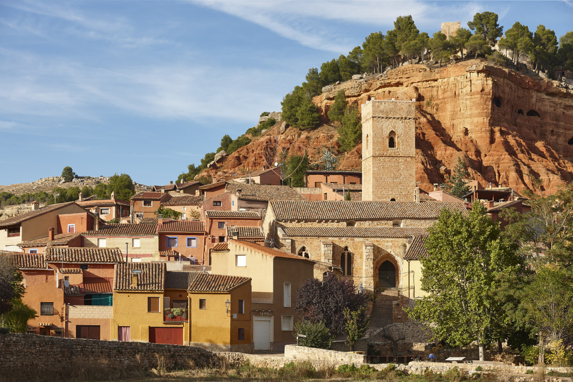 The stone Church of San Blas stands among terracotta-roofed houses in Anento, Zaragoza, Spain. Red sandstone cliffs, pine trees and the remains of a hilltop castle rise behind the village.