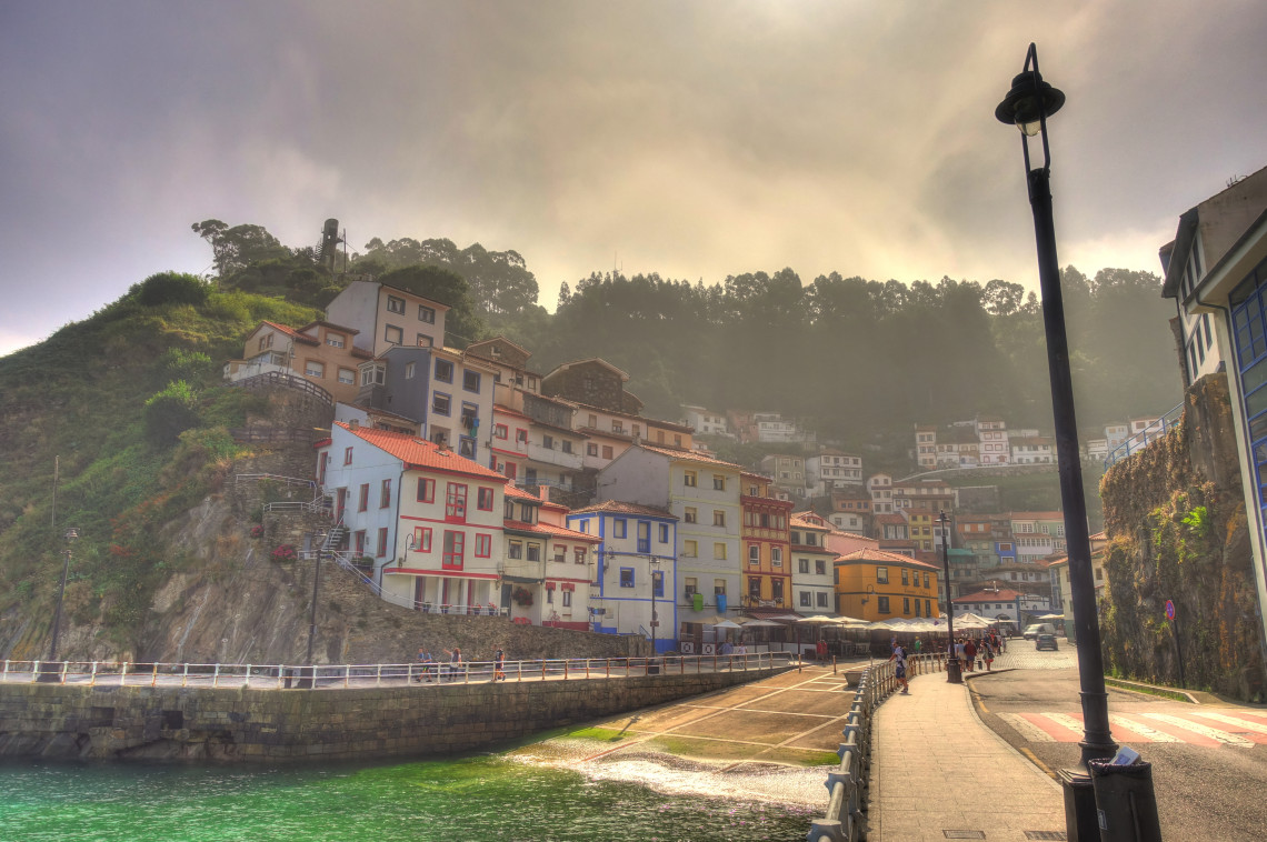 Colourful houses climb the steep hillside above the old fishing port of Cudillero in Asturias, Spain. A waterfront promenade follows the sheltered harbour beneath misty, wooded slopes.