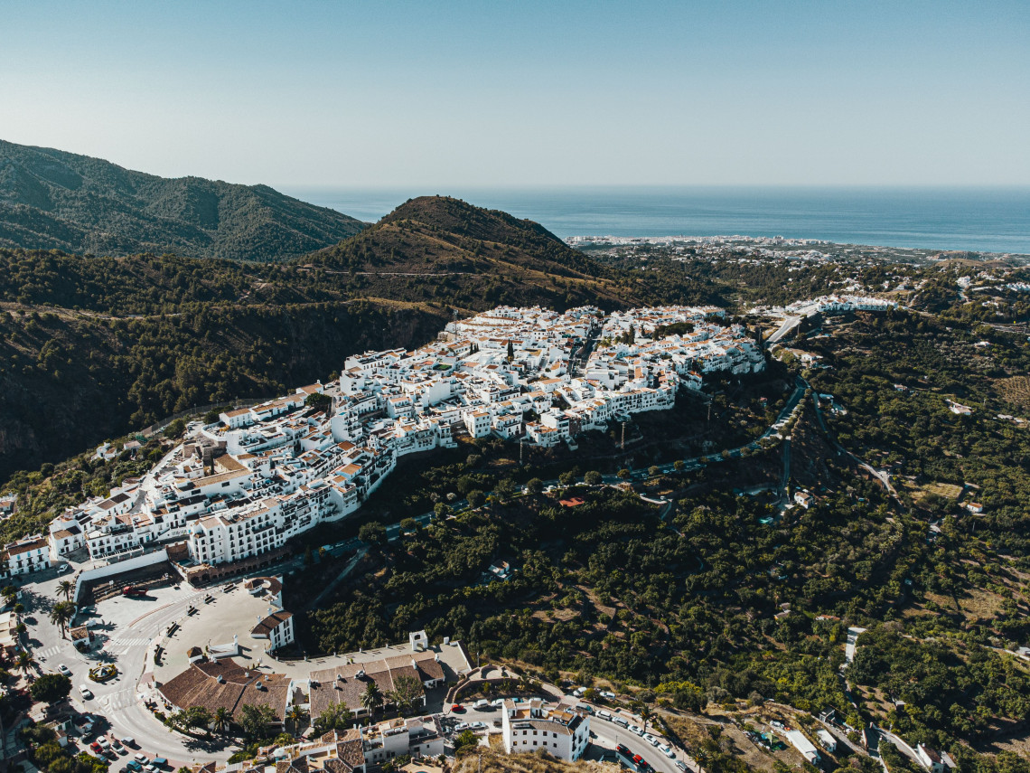 An aerial view shows the whitewashed buildings and terracotta roofs of Frigiliana in M&aacute;laga, Spain, clustered across a hillside. Forested mountains surround the village, with the Mediterranean Sea visible beyond the coastal plain.
