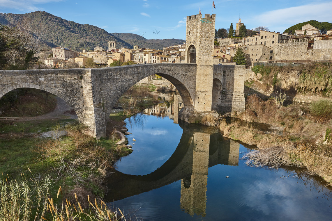 The medieval stone bridge of Besal&uacute; crosses the Fluvi&agrave; River in Girona, Spain. Its arched spans and fortified gateway tower are reflected in the calm water below the historic town.