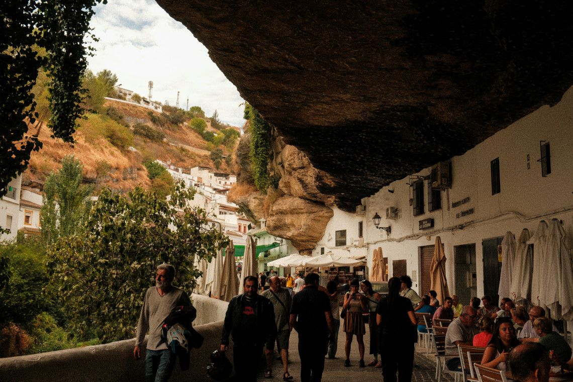Restaurants and whitewashed buildings occupy the space beneath a large natural rock overhang on Cuevas del Sol in Setenil de las Bodegas, C&aacute;diz, Spain. Visitors walk along the shaded cliffside street beside outdoor caf&eacute; tables.
